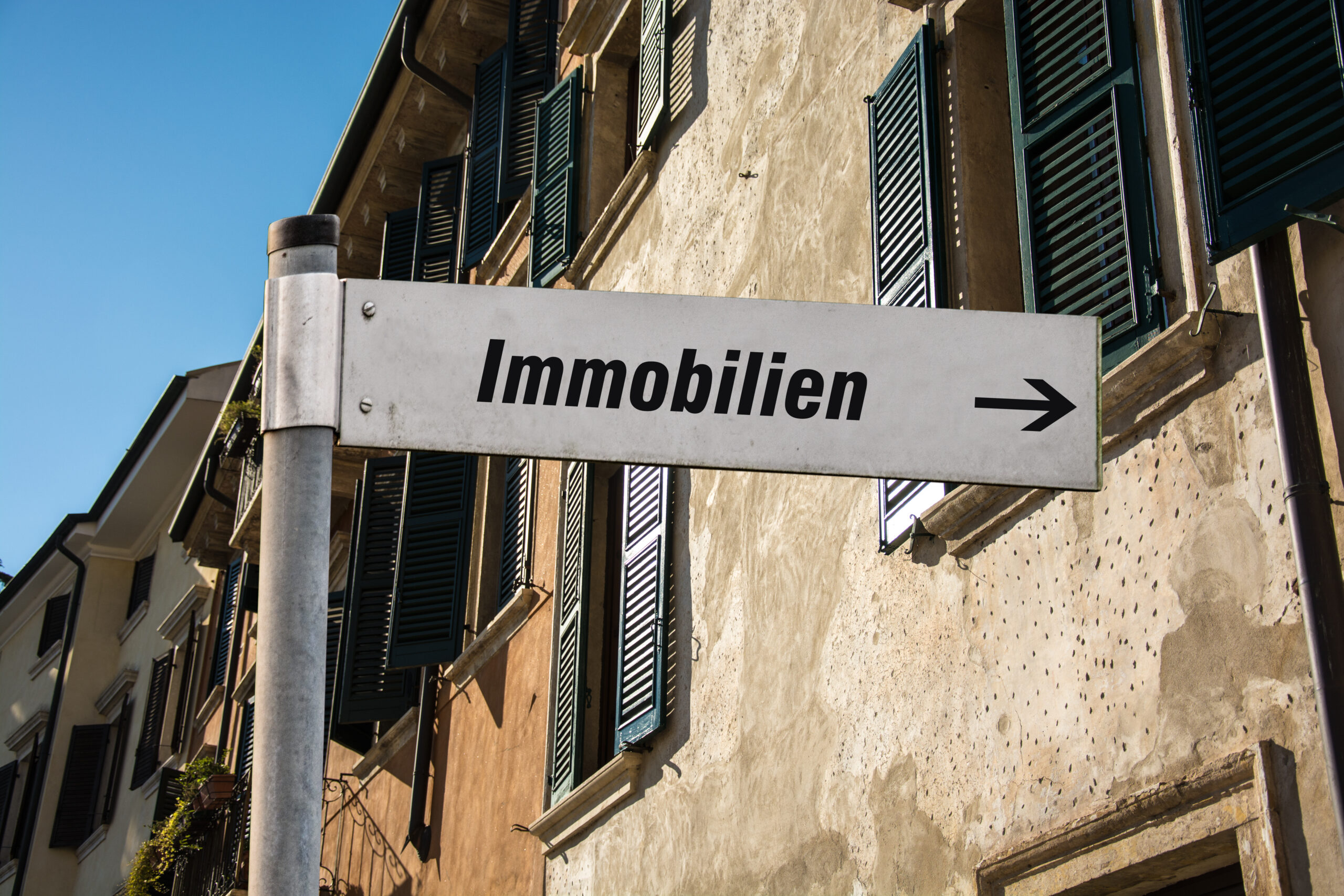 Schild mit der Aufschrift "Immobilien" zeigt nach rechts. Hintergrund: Gebäude mit grünen Fensterläden.