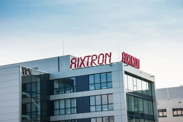 Glasgebäude mit rotem AIXTRON Schriftzug auf dem Dach unter blauem Himmel.