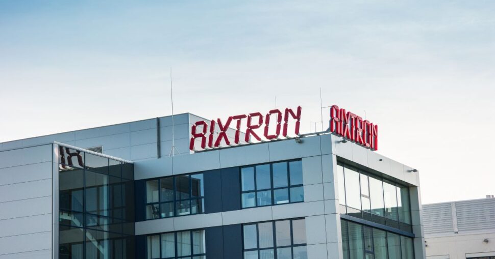 Glasgebäude mit rotem AIXTRON Schriftzug auf dem Dach unter blauem Himmel.
