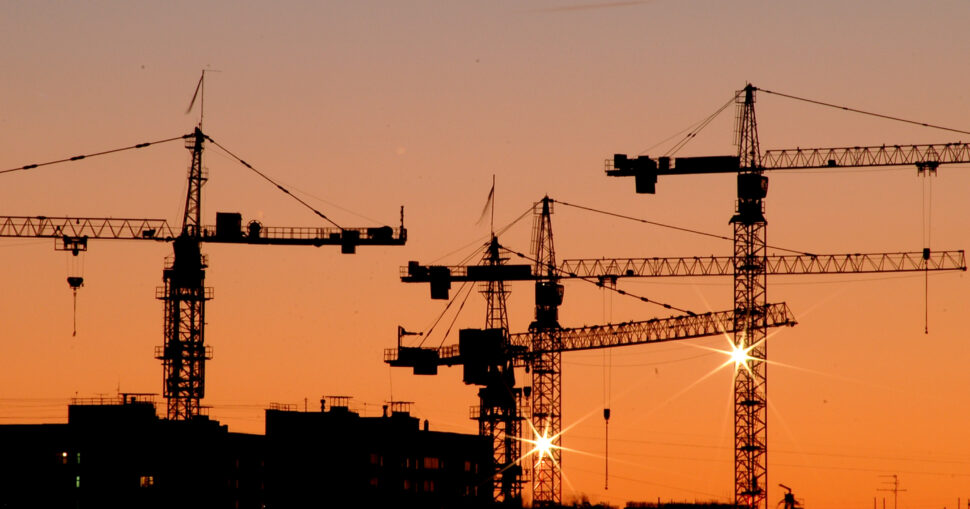 Kräne vor einem orangefarbenen Himmel im Sonnenuntergang. Silhouetten von Gebäuden im Vordergrund.