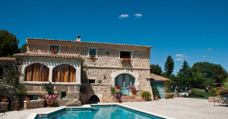 Steinhaus mit Blumen, Pool im Vordergrund, blauer Himmel, Bäume im Hintergrund.