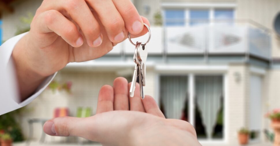 Eine Hand übergibt Schlüssel an eine andere Hand vor einem Haus im Hintergrund.